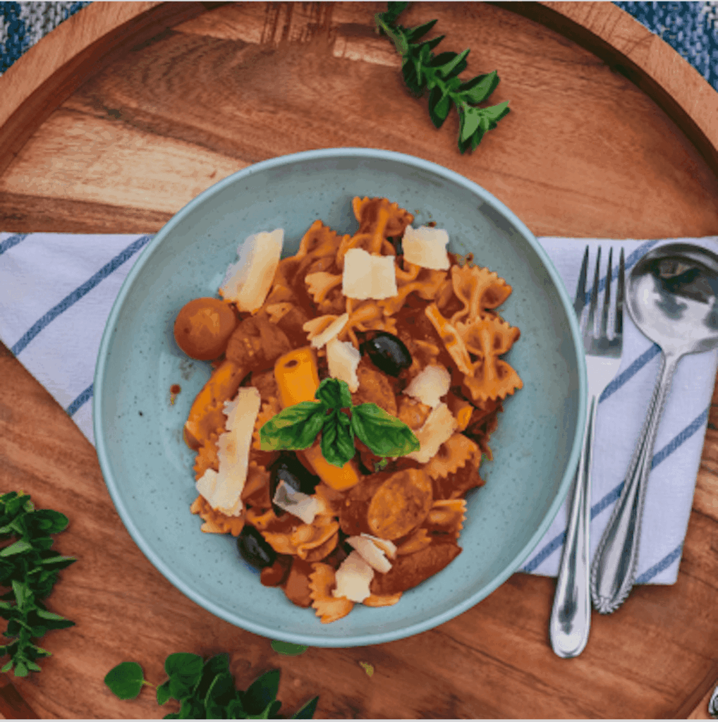 Bowl of pasta with sliced sausage, peppers, tomato sauce, and shaved cheese.
