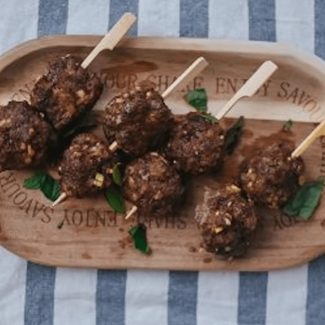 Thai Inspired Meatballs Cooked meatballs on skewers served on a wooden board with herbs.